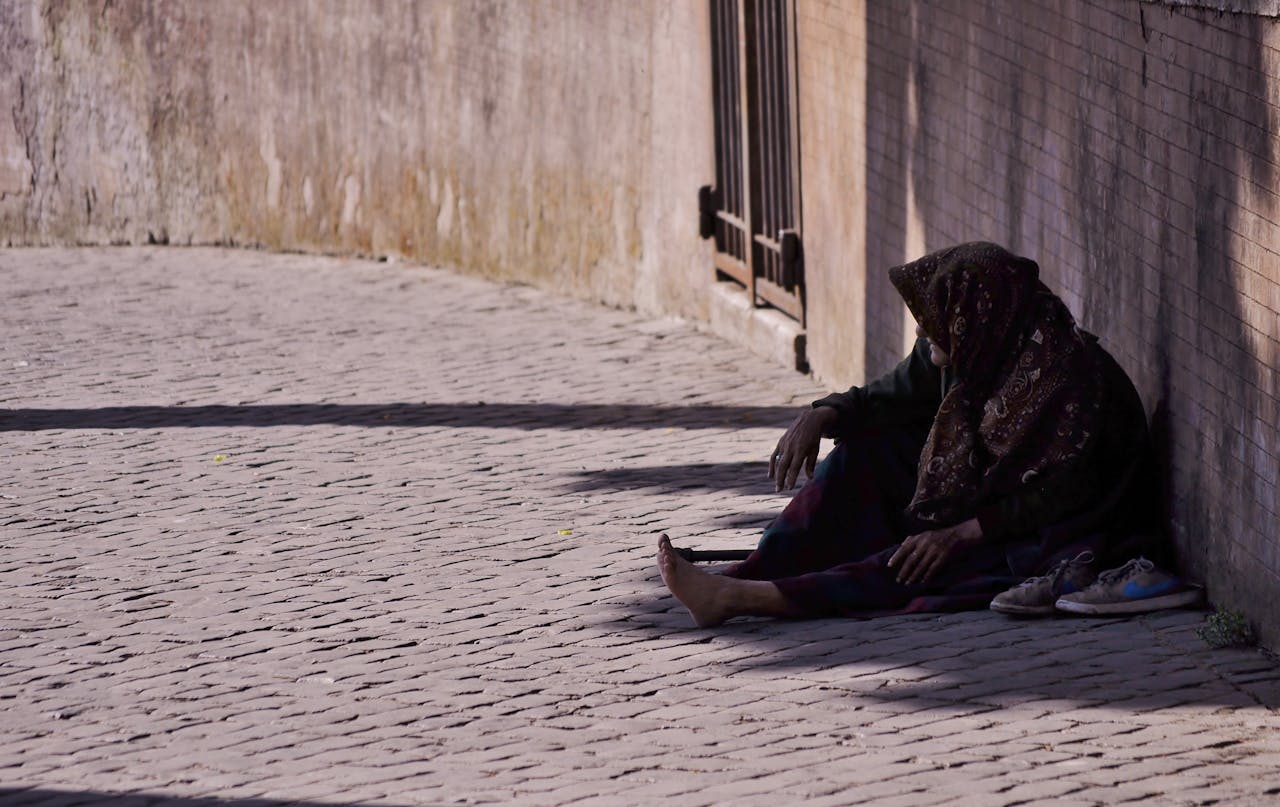 who-we-are A woman sitting alone on a cobblestone street, covered with a headscarf.