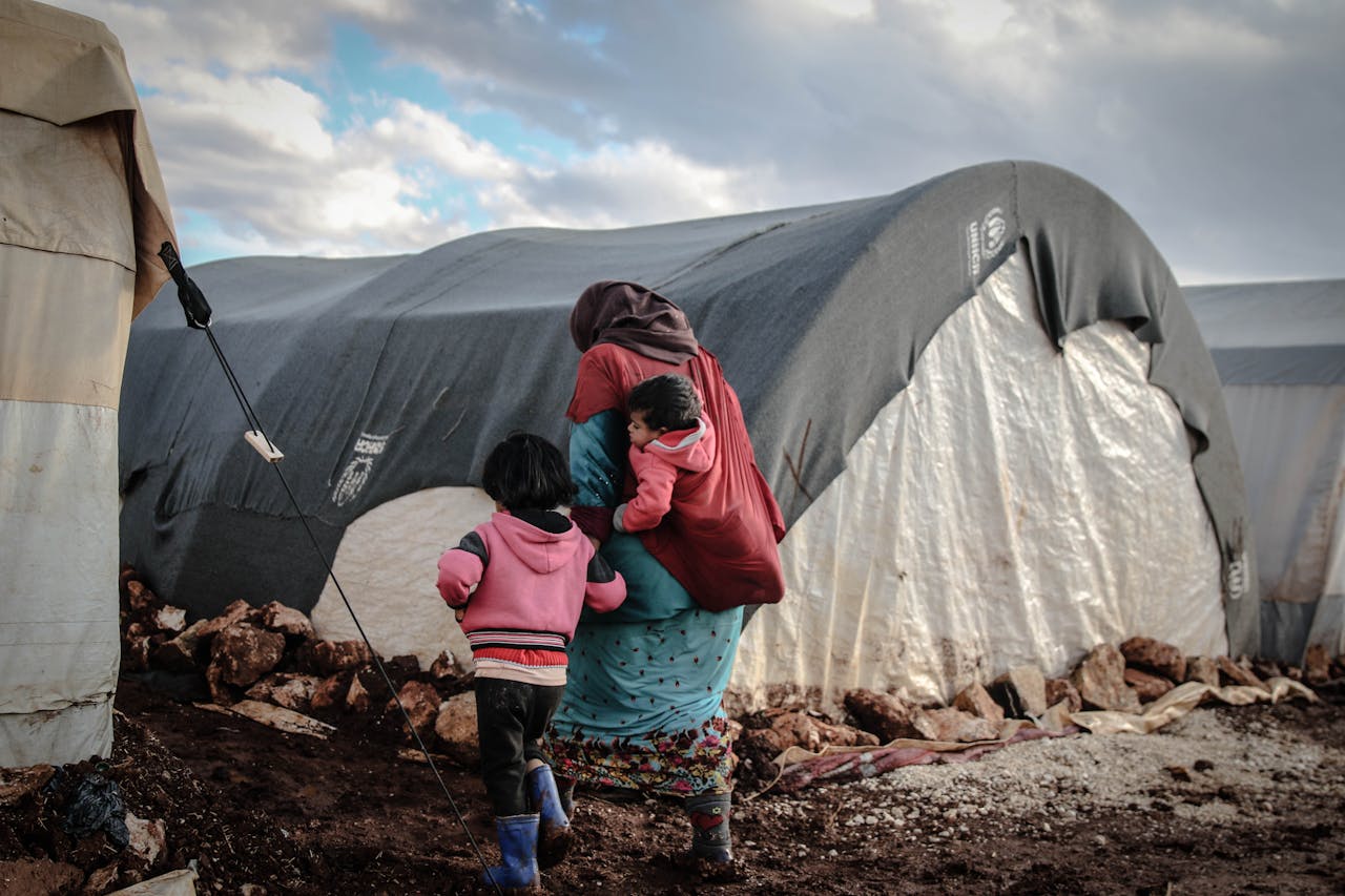 services-04 A woman and children walk near tents in a refugee camp in Idlib, showcasing resilience amidst adversity.