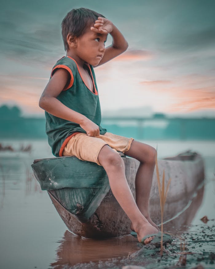 services-02 A young boy sits on a wooden boat by a misty riverside in Nepal, gazing into the distance.